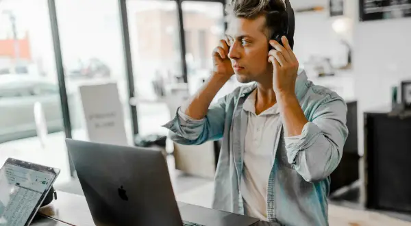 A young man, likely one of the new generation of music managers, adjusts his headphones while working on his laptop in a cafe.