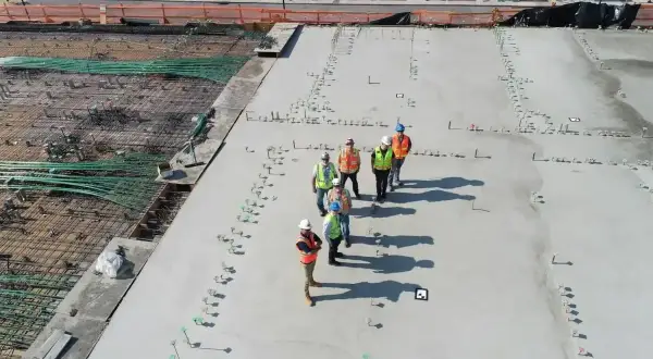 Grupa budowlańców na placu budowy, dyskutująca o cenie betonu i postępach prac. W tle zbrojenie i świeżo wylany beton.