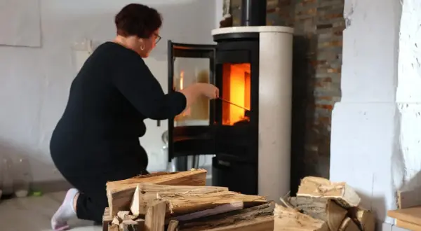 Frau kniet vor einem brennenden Kamin, um ihn zu reinigen. Holzscheite liegen bereit.