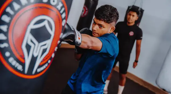 A young boxer practices his boxing combos, throwing a punch at a heavy bag in a gym. Another person watches in the background.
