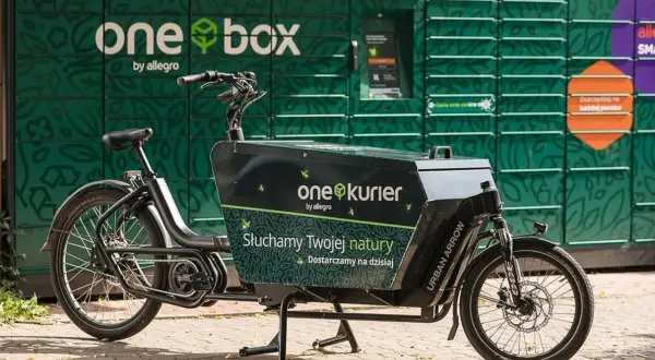 Rower cargo z napisem "one kurier by allegro" stoi przed zielonym paczkomatem "one box".