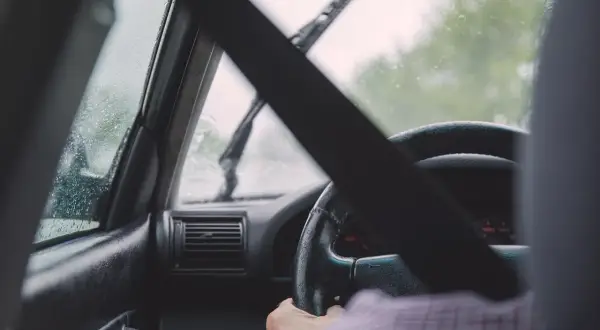 Widok z wnętrza samochodu podczas jazdy w deszczu. Krople na szybie, wycieraczki pracują. Kierowca trzyma kierownicę.