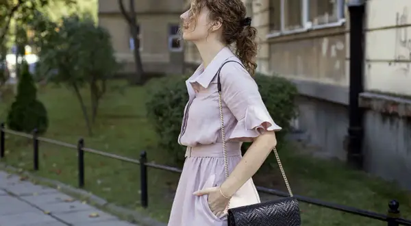 Kobieta w różowej sukience i czarnej torebce na łańcuszku na tle budynku i zieleni.