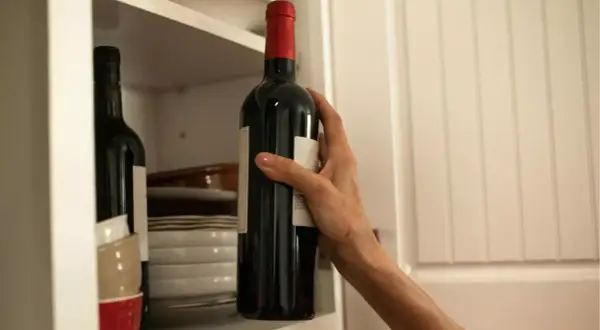 Dłoń sięga po butelkę bordeaux wina z półki, obok stosu talerzy i miseczek.