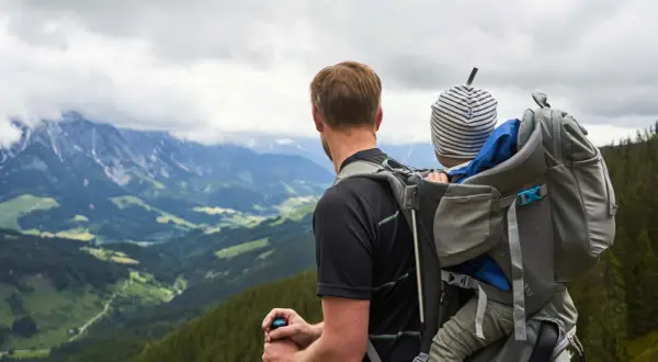 Mann mit Kind auf dem Rücken in einer Kraxe, blickt auf bergige Landschaft.