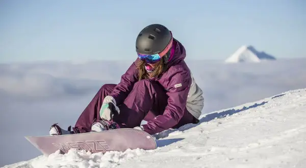 Kobieta w kasku i goglach siedzi na śniegu, zapinając deskę snowboardową.