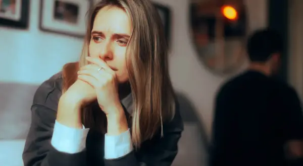 Zamyślona kobieta z dłońmi przy ustach, obok niewyraźna postać mężczyzny. Czuć dystans w związku.