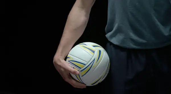 Dłoń zawodnika trzyma piłkę do siatkówki. Ciemne tło podkreśla biało-żółto-niebieską piłkę, gotową do gry w siatkówkę mężczyzn plusliga.