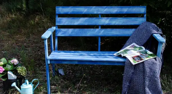 Niebieska ławka ogrodowa z kocem i czasopismem, obok konewka i kwiaty.