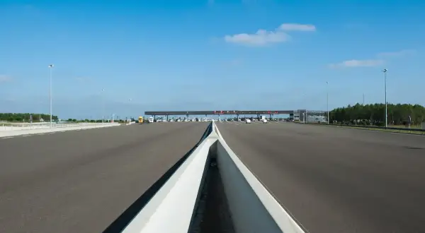 Péage autoroutier sous un ciel bleu. La route s'étend vers une barrière de péage avec plusieurs voies. Un camion jaune est visible.