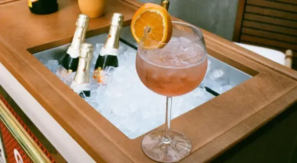 Orzeźwiający drink z prosecco i colą, ozdobiony plasterkiem pomarańczy, obok schłodzonych butelek.