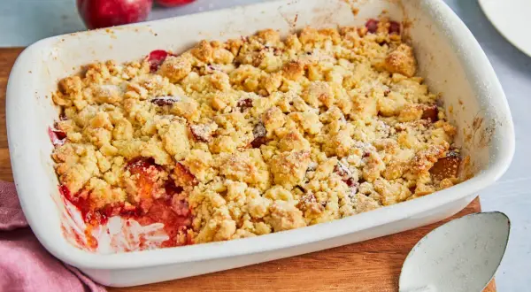 Ein frisch gebackener pflaumen crumble in einer weißen Auflaufform, bereit zum Servieren. Daneben liegen zwei reife Pflaumen.