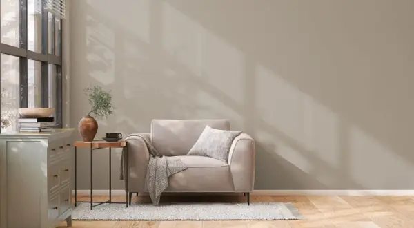 Un sillón beige con manta y cojín, junto a una mesita auxiliar con jarrón y taza. La pared tiene unos colores de pintura neutros, con sombras de luz.