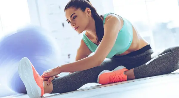Femme en tenue de sport faisant un exercice souplesse, étirant sa jambe droite. Une grosse balle de gym est visible en arrière-plan.
