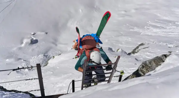 Escalade d'une échelle sur un flanc de montagne enneigé, skis sur le dos. L'ascension vers un sommet 4000 des Alpes, une aventure pas si facile.