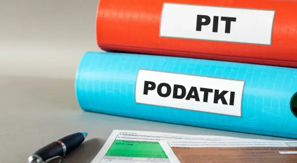 Dwa segregatory, jeden czerwony z napisem "PIT", drugi niebieski z napisem "PODATKI", leżą na biurku obok formularzy podatkowych i długopisu.