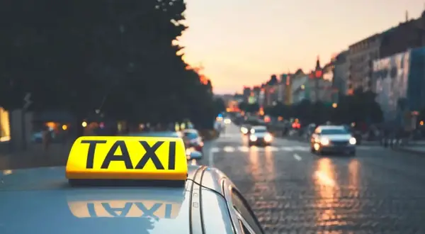 Żółty znak TAXI na dachu samochodu na tle miasta o zachodzie słońca.