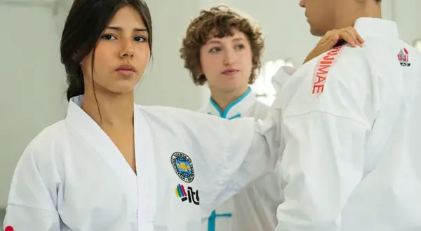 Trójka młodych ludzi w białych strojach do taekwondo, jednej z najpopularniejszych sztuk walki, ćwiczy w sali treningowej.