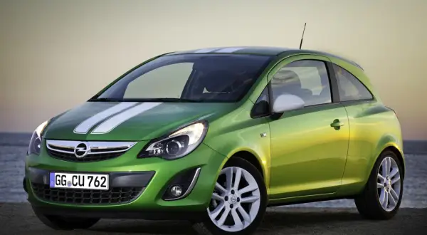 Zielony Opel Corsa D z białymi pasami na masce i dachu, zaparkowany na plaży o zachodzie słońca.