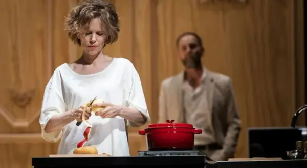 La locandiera recensioni: donna sbuccia una mela, pentola rossa sul fuoco, uomo sullo sfondo.