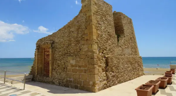 Antica torre di pietra sulla costa, con il mare azzurro sullo sfondo. Vicino, una fila di vasi di terracotta, come tre fontane in attesa.