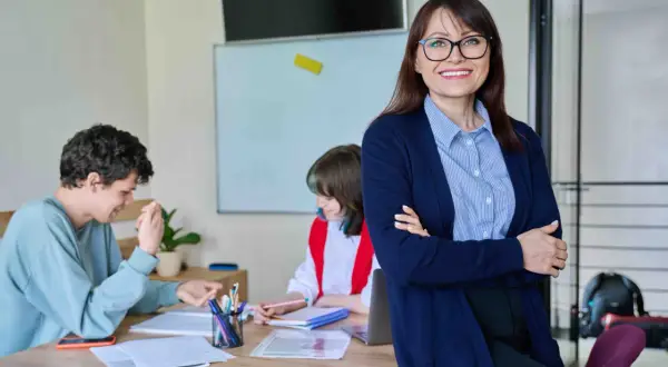 Nauczycielka z uśmiechem nadzoruje zajęcia z wychowawcą. Uczniowie pracują przy biurku, a na tablicy widać rozkład materiału.
