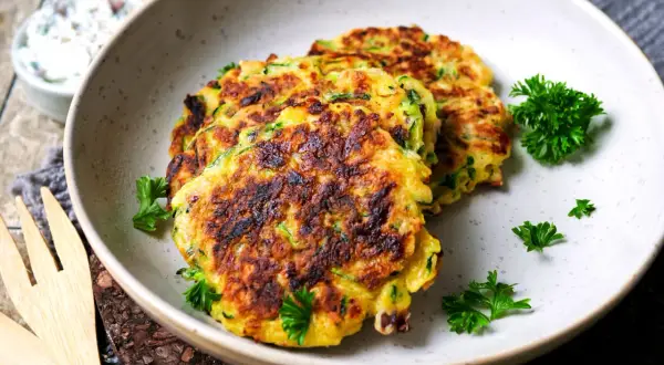 Goldbraune Zucchini-Puffer, garniert mit Petersilie, liegen auf einem Teller. Daneben eine Schüssel mit Dip.