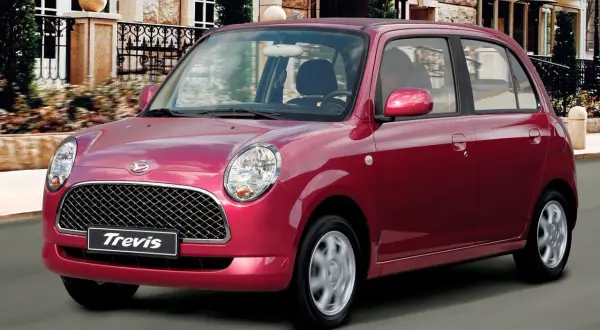 Czerwony Daihatsu Trevis z charakterystycznym grillem i okrągłymi światłami jedzie ulicą przed eleganckim budynkiem.
