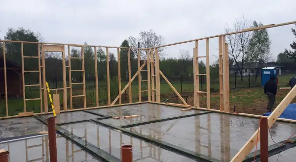 Budowa fundamentu pod dom szkieletowy. Drewniana konstrukcja ścian stoi na betonowej płycie, widać rury kanalizacyjne.