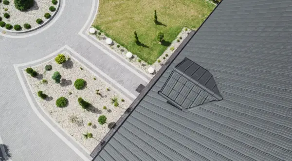Nowoczesny dach pulpitowy z szarej blachy, z widocznym oknem dachowym. Obok zadbany ogród z brukowanymi ścieżkami i niską roślinnością.