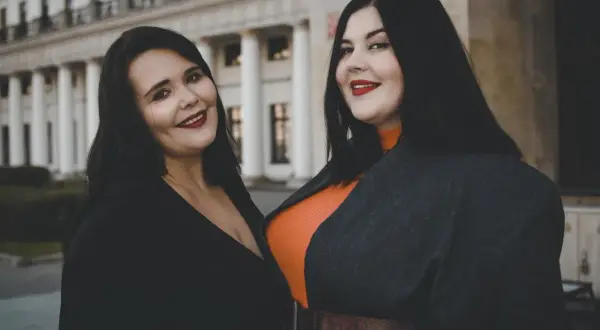 Dwie uśmiechnięte kobiety, uosobienie body positivity, pozują przed budynkiem z kolumnami.