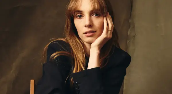Maya Hawke w czarnej marynarce, opierająca głowę na dłoni. Aktorka znana z filmów, seriali i programów.