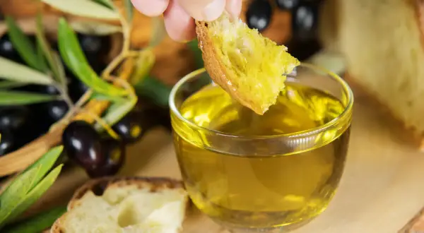 Un pezzo di pane intinto in olio dorato, con olive e rami d'ulivo sullo sfondo, evoca le antiche origini olio di oliva.