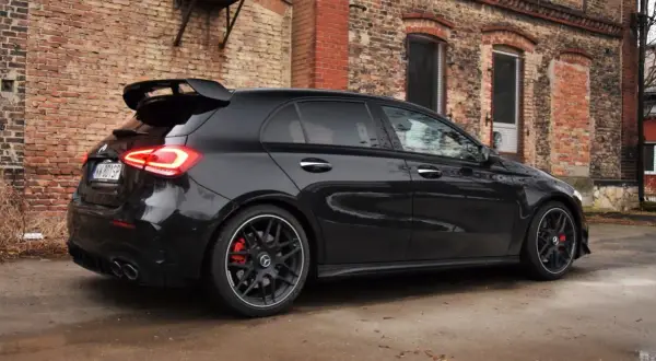 Czarny Mercedes-AMG A45s z masywnym spojlerem i czerwonymi zaciskami hamulcowymi stoi na mokrej nawierzchni przed starą ceglaną kamienicą.