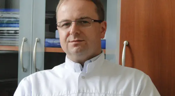 Portret mężczyzny w okularach i białym fartuchu lekarskim, na tle regałów z książkami.