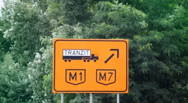 Pomarańczowy znak drogowy w drodze na Węgry. Informuje o tranzycie ciężarówek na autostrady M1 i M7.
