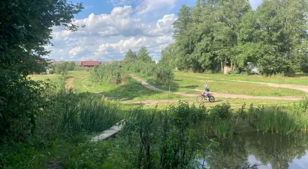 Osoba na motocyklu crossowym jedzie po szutrowej drodze wśród zieleni, obok stawu i drewnianego mostku.