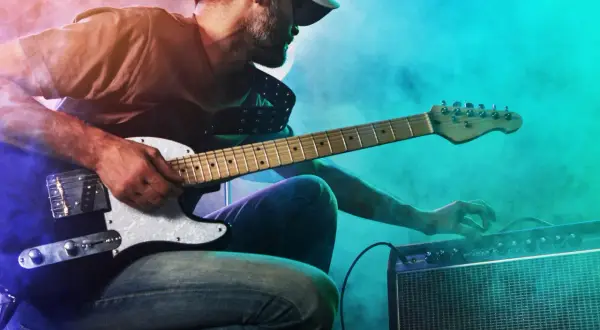Mann mit Gitarre und Verstärker, der den perfekten gitarren sound einstellt. Nebel und bunte Lichter schaffen eine rockige Atmosphäre.