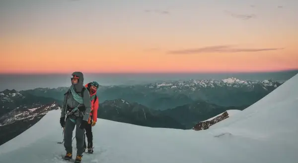 Deux alpinistes entament leur ascension du Mont Rose au lever du soleil, le ciel aux teintes roses et orangées embrassant les sommets enneigés.