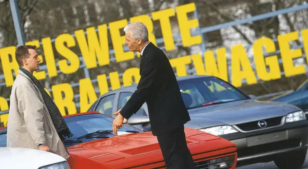 Dwóch mężczyzn ogląda sportowe auta. Jeden wskazuje na czerwony samochód, drugi stoi obok.