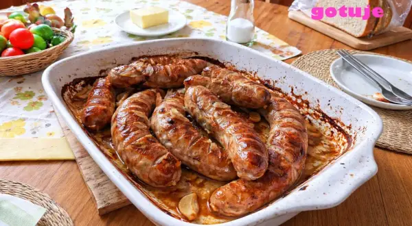 Pyszna, pieczona kiełbasa biała w naczyniu żaroodpornym, z cebulką i czosnkiem. Obok koszyk z jajkami i kromka chleba.