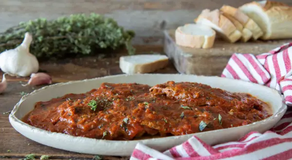 Herzhaftes Carne alla Pizzaiola in Tomatensauce, garniert mit Kräutern, neben Knoblauch, Brot und einem rot-weiß gestreiften Tuch.