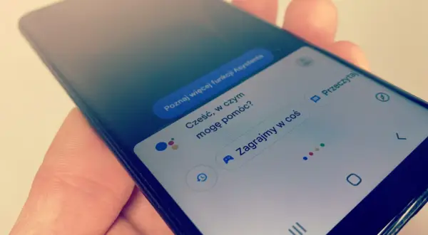 Smartfon z Asystentem Google, który czeka na **wyszukiwanie głosowe**. Widoczne opcje: "Poznaj więcej funkcji Asystenta", "Cześć, w czym mogę pomóc?", "Zagrajmy w coś".