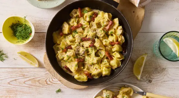Pyszne tortellini grzybowe w kremowym sosie, posypane parmezanem i boczkiem, podane z cytryną i koperkiem.