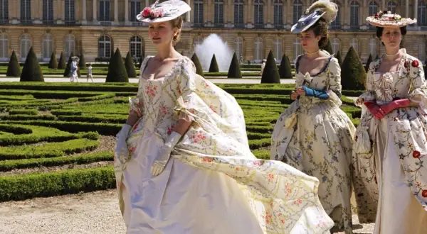 Kvinnor i magnifika **rococo fashion**-klänningar med blomstermönster promenerar i en formell trädgård framför ett palats.