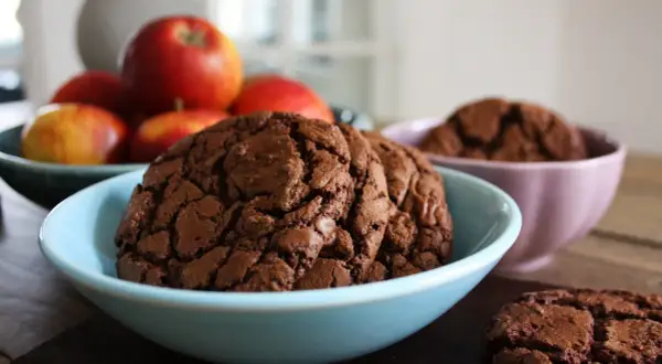Mörka, sega kladdkake cookies i en ljusblå skål, med äpplen i bakgrunden. En annan skål med cookies syns i bakgrunden.