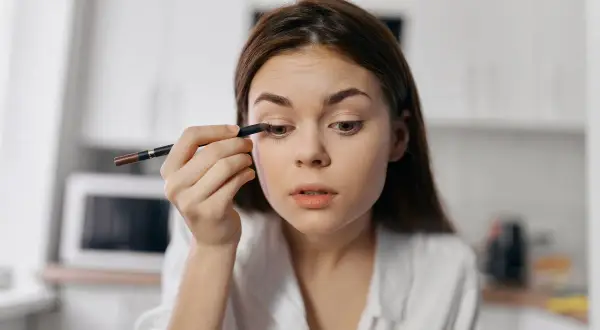 Dziewczyna wykonuje subtelny makijaż typu clean girl, precyzyjnie podkreślając oko kredką.