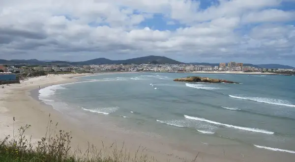 Amplia vista de la playa de Altar Barreiros con arena dorada, mar azul y una ciudad costera al fondo.