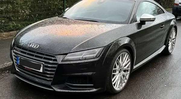 Negro Audi TT RS mojado por la lluvia, con reflejos naranjas en el capó. El coche está listo para la etapa 1 de su viaje.