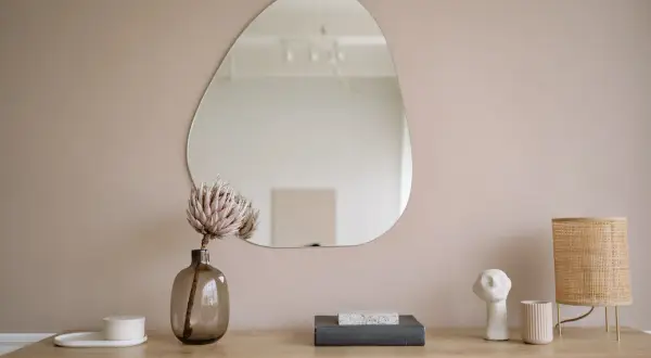 Un miroir aux formes organiques reflète la pièce. Sur le meuble en bois, une fleur séchée, des livres, une lampe en rotin et des objets décoratifs créent une scène apaisante.